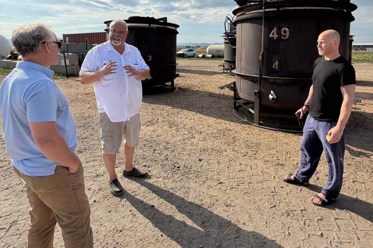Three men talking on a gravel yard beside large steel biochar kilns under a cloudy sky.